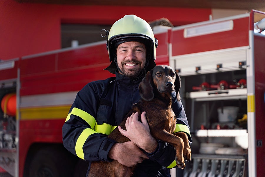 Firefighter in uniform and helmet smiles while holding a brown dachshund in front of a fire truck.