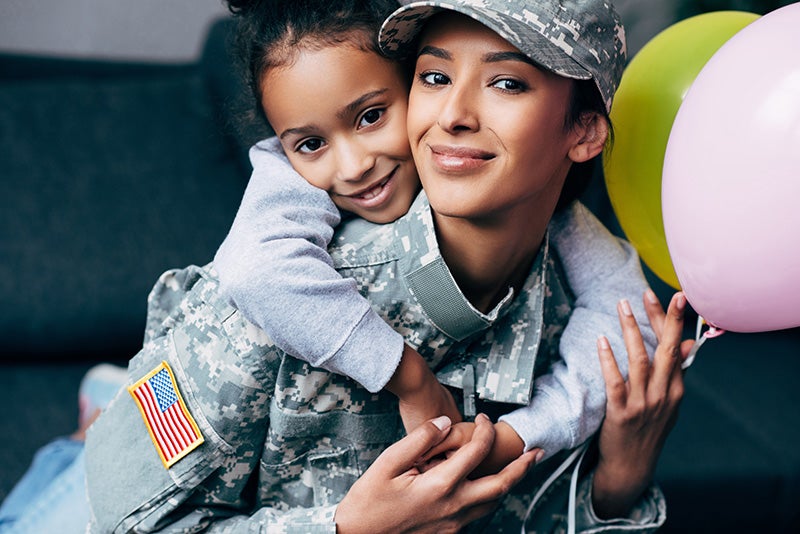 A smiling young girl hugs a female soldier in camouflage uniform, who also smiles and wears a cap.