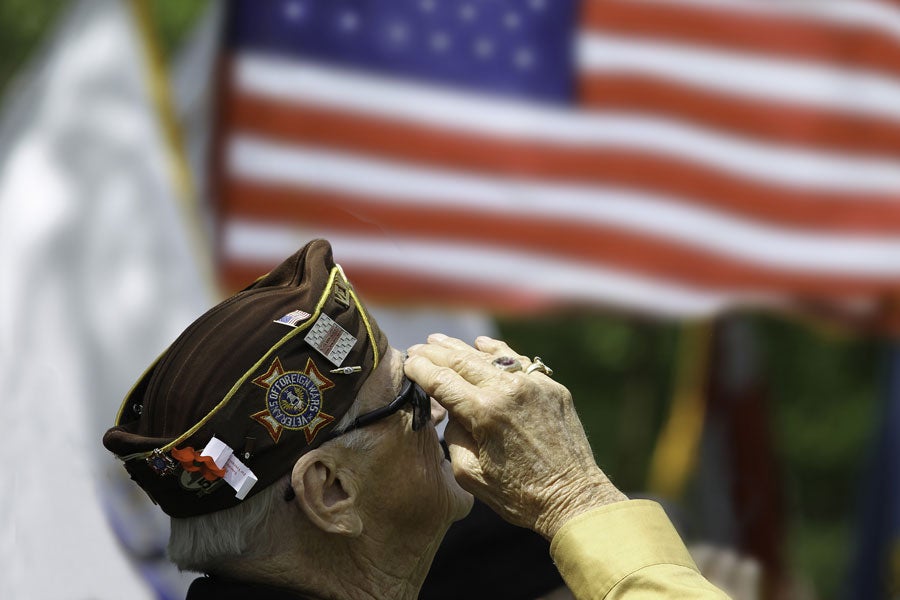 An elderly veteran in a brown VFW cap salutes, with a blurry American flag in the background.