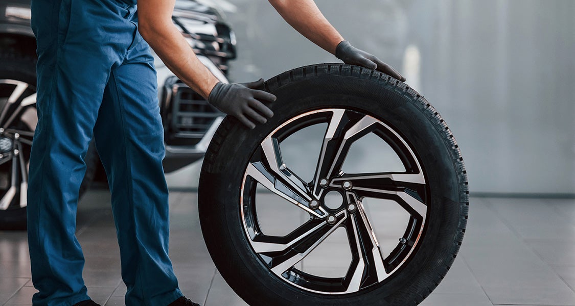 A mechanic in blue overalls and gloves rolls a car tire with a black and silver rim across a garage floor.